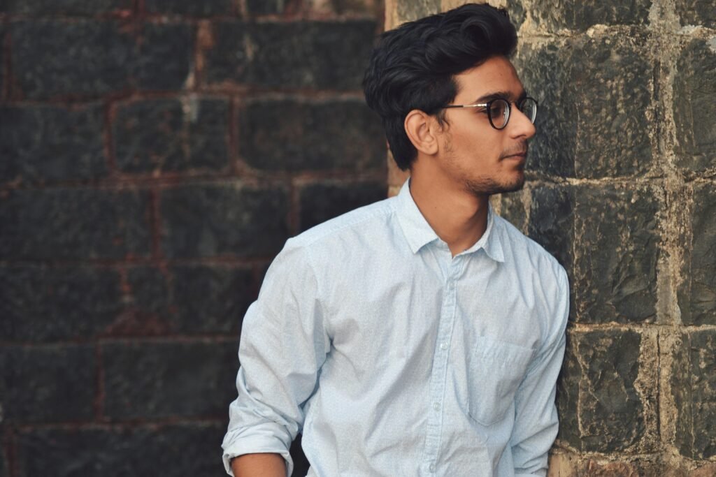 Home A stylish young man in glasses leans against a brick wall wearing a casual light blue shirt.